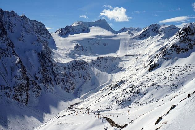 Une vaste chaîne de montagnes enneigée avec des remontées mécaniques et des pistes visibles sous un ciel bleu éclatant avec quelques nuages.