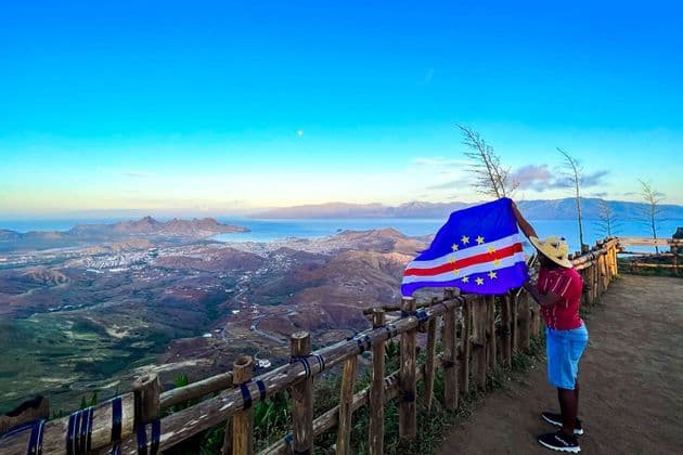 Eine Person mit Hut hält die Flagge Kap Verdes und blickt von einem Aussichtspunkt über eine Küstenstadt und Berge.