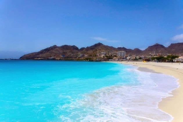 Türkisfarbene Wellen spülen an einen breiten Sandstrand, mit einer Küstenstadt und trockenen Bergen im Hintergrund unter einem klaren blauen Himmel.