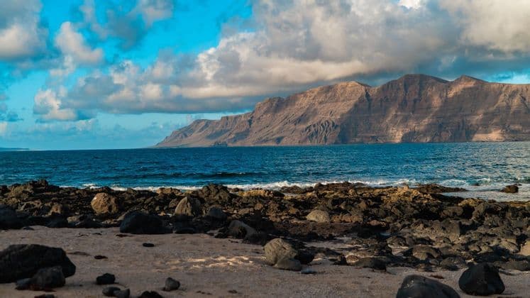 Una costa rocosa con arena y piedras volcánicas se enfrenta a una gran cordillera al otro lado del océano azul bajo un cielo parcialmente nublado.