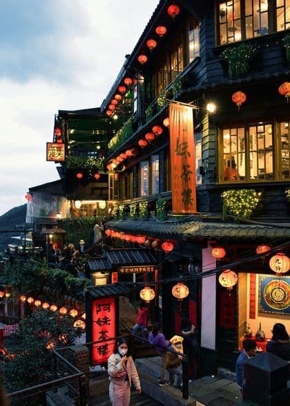 Al crepuscolo, le persone percorrono le scale tra edifici tradizionali collinari, illuminati da numerose lanterne di carta rosse e luminose.