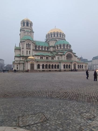 Eine große, weiße Kathedrale mit goldenen Kuppeln und grünen Dächern steht an einem nebligen Tag auf einem weitläufigen Kopfsteinpflasterplatz.