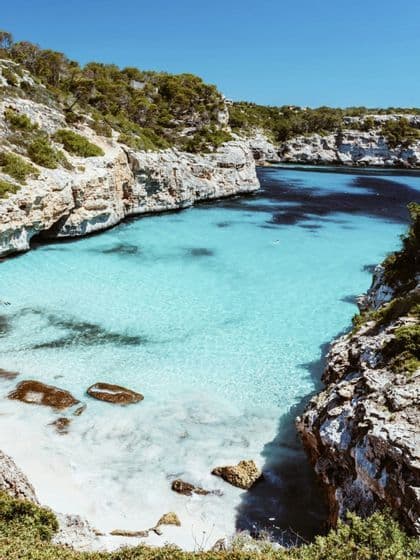 Una vista da un'alta scogliera di una caletta appartata con acqua turchese trasparente, una piccola spiaggia di sabbia bianca e coste rocciose.