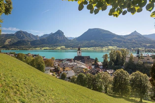 Una vista da una collina erbosa di un villaggio con un campanile di chiesa sulla riva di un lago turchese, con grandi montagne sullo sfondo.
