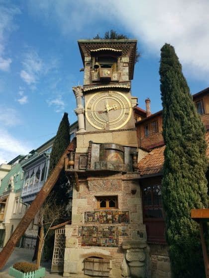 A whimsical, leaning clock tower with a large golden face stands on a city street next to a tall cypress tree.