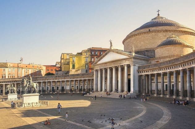 Une vaste place ensoleillée où des gens se promènent devant un grand bâtiment à dôme et à colonnade classique.