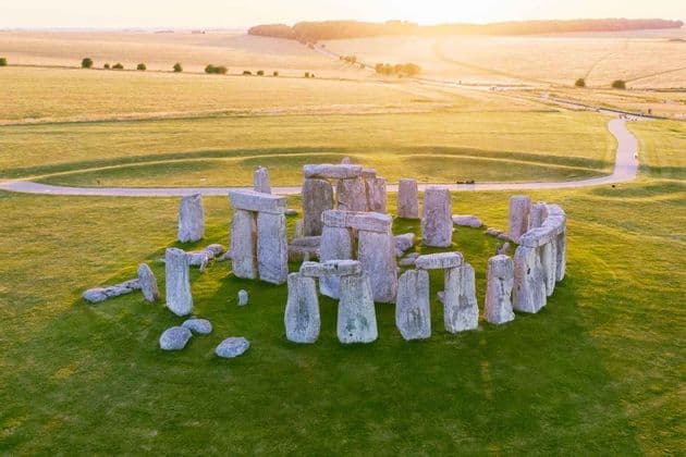 Una vista aerea del monumento di Stonehenge su un prato verde, illuminato dalla luce dorata del tramonto su campi ondulati.