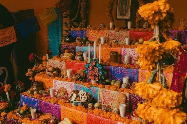 A colorful, multi-tiered Day of the Dead altar decorated with orange marigolds, candles, fruit, bread, and skulls.