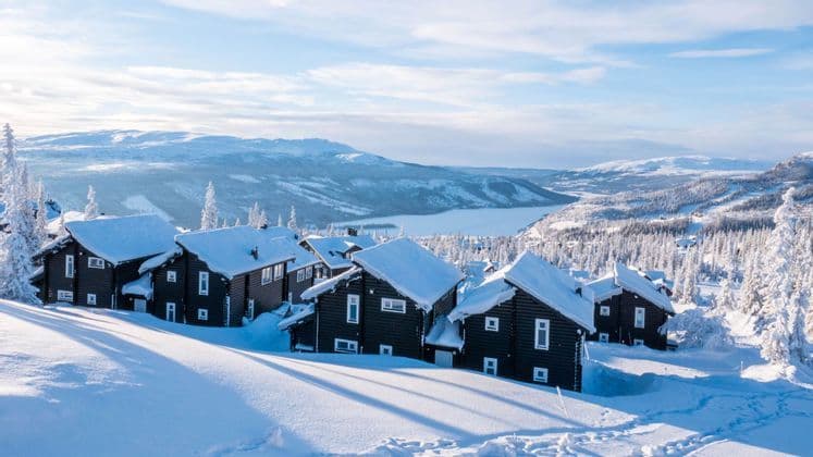 Una fila di baite in legno scuro con tetti coperti di neve si trova su una collina soleggiata e innevata che domina un vasto paesaggio invernale.
