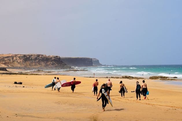 Un groupe WeRoad en combinaisons de surf, planches à la main, marche sur une plage de sable vers l'océan, avec des falaises au loin.