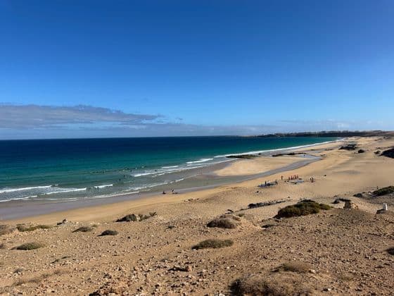 Un voyage de groupe WeRoad avec une leçon de surf sur une vaste plage de sable aux eaux turquoise, et des surfeurs dans les vagues.