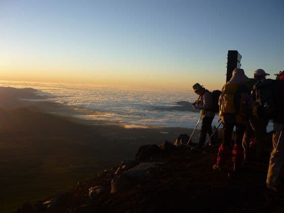 Un gruppo WeRoad di escursionisti con zaini si erge su una cima rocciosa all'alba, ammirando un vasto mare di nuvole.