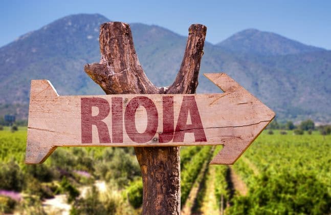 Un panneau fléché en bois indiquant 'Rioja' pointe vers la droite, avec un vignoble et des montagnes en arrière-plan.