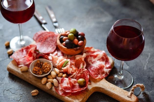 Une planche de charcuterie en bois avec des viandes salées, des olives et des noix est servie avec deux verres de vin rouge.