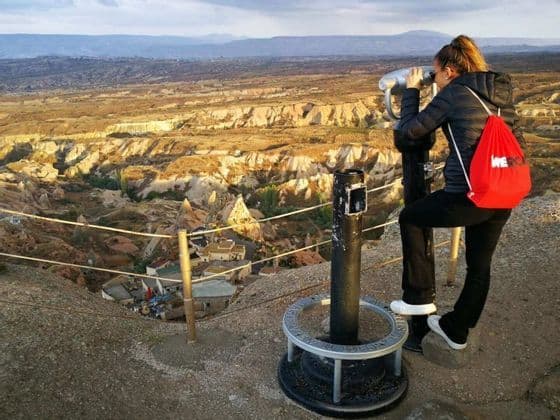 Una mujer con una mochila roja WeRoad mira a través de un visor, contemplando un vasto valle rocoso desde un mirador elevado.