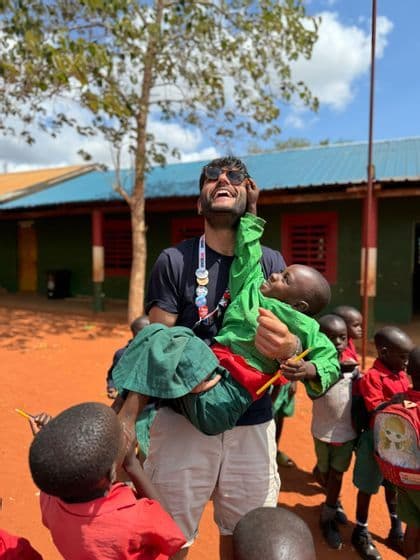 Un uomo in viaggio di gruppo WeRoad sorride e solleva un bambino in uniforme verde, circondato da altri bambini nel cortile di una scuola.