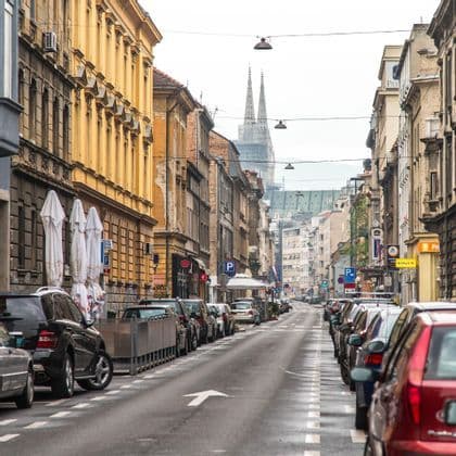 A long city street lined with parked cars and historic buildings, with two cathedral spires visible in the distance under an overcast sky.