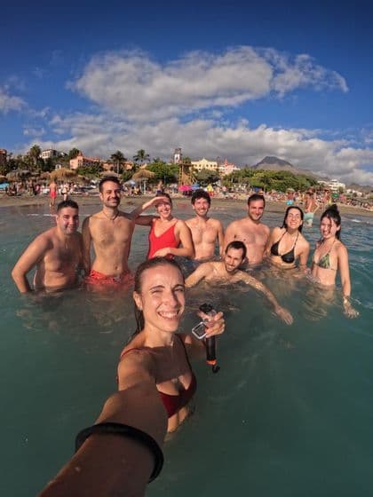 Eine Frau macht ein Selfie mit ihrer WeRoad-Gruppe, während sie an einem überfüllten Strand unter teilweise bewölktem Himmel im Meer schwimmt.