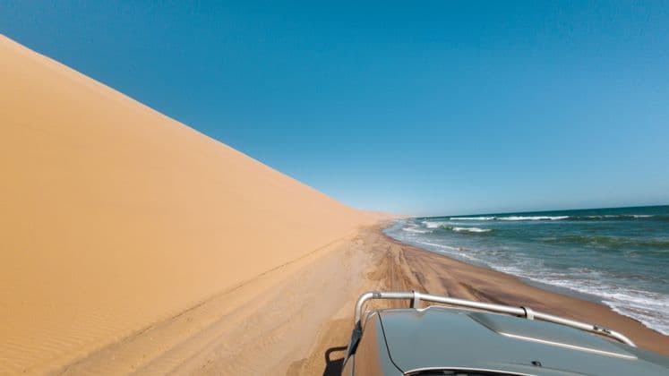 Ein Blick aus einem Geländewagen auf einem Sandweg zwischen einer großen Sanddüne und den Meereswellen unter klarem Himmel.