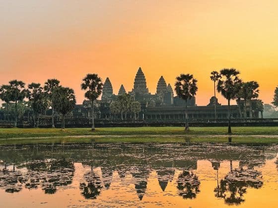 An ancient temple with tall spires and palm trees reflected in a calm body of water against an orange sky at sunrise.
