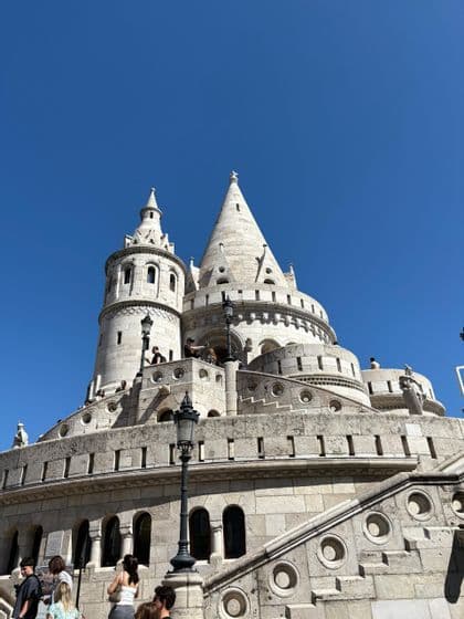 Vue en contre-plongée d'un bastion en pierre blanche avec des tours coniques, avec des gens sur ses escaliers et balcons sous un ciel bleu clair.