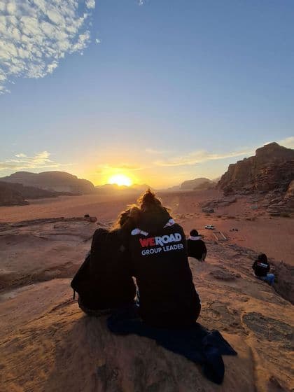 Eine WeRoad-Gruppe mit Reiseleiter sitzt auf einem felsigen Hügel und beobachtet den Sonnenuntergang über einer Wüstenlandschaft.