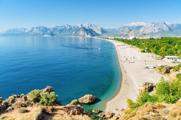 A panoramic view of a long sandy beach along a calm blue sea, with a coastal city and mountains in the background.