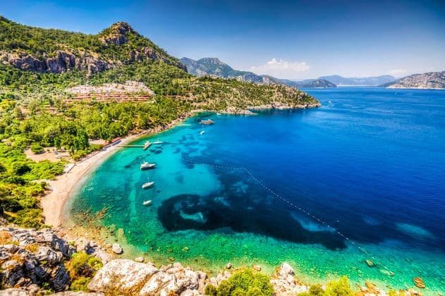 An aerial view of a bay with clear turquoise water, a sandy beach, and boats anchored along a mountainous coast covered in lush green trees.