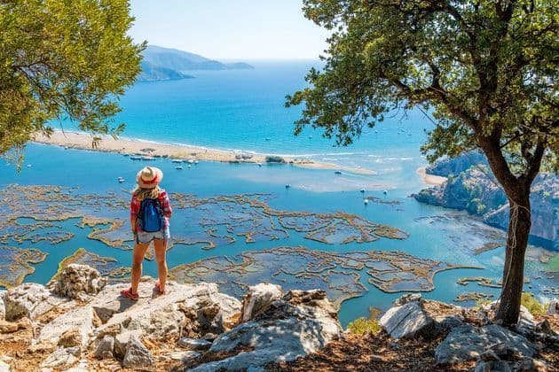 A person with a backpack stands on a rocky viewpoint, looking out at an expansive coastal delta and the sea beyond.