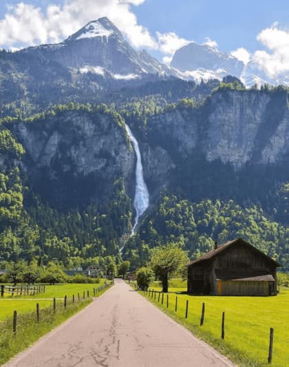 A paved road through a green valley leads to a tall waterfall cascading down a forested mountain with snow-capped peaks under a blue sky.