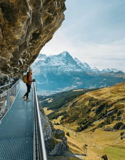 A woman with a backpack stands on a metal walkway attached to a cliff, overlooking a vast mountain valley with snow-capped peaks.