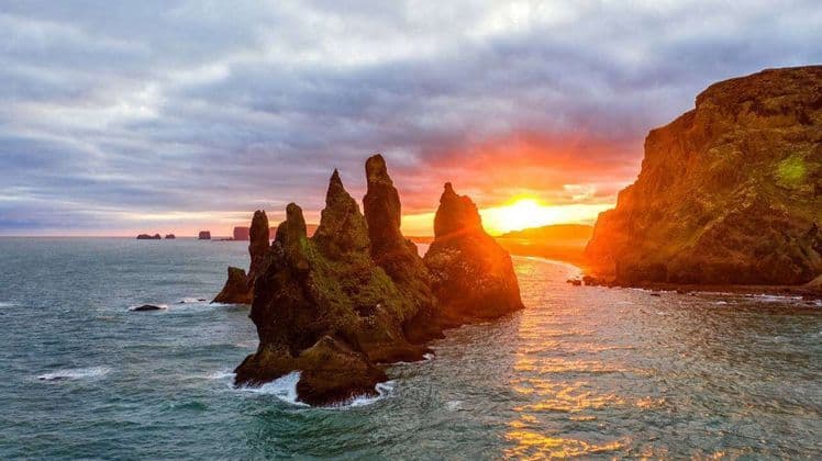 Zackige, moosbewachsene Felsnadeln ragen aus dem Meer, während die Sonne untergeht und ein orangefarbenes Glühen auf das Wasser wirft.