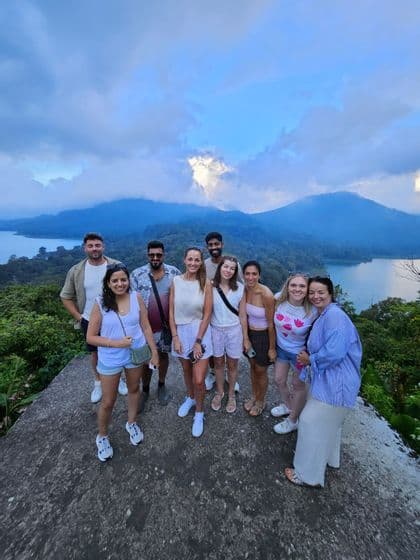 Un groupe WeRoad de neuf personnes posant sur un belvédère surplombant un lac et des montagnes verdoyantes sous un ciel nuageux.