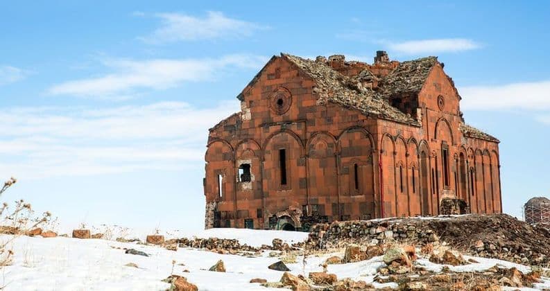 Una cattedrale in pietra rossa in rovina con il tetto crollato si erge su una collina innevata sotto un cielo azzurro parzialmente nuvoloso.