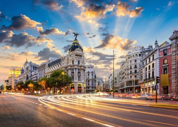 A long exposure shot of a wide city street at sunset, with light trails from moving traffic and historic buildings under a dramatic sky.