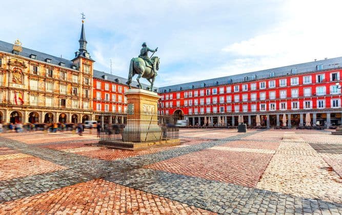 A bronze equestrian statue stands in the center of a large cobblestone plaza, surrounded by historic red buildings as people walk by.