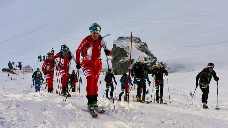 Un gruppo di sci alpinisti in tenuta completa procede in fila in salita su una montagna innevata durante una gara.