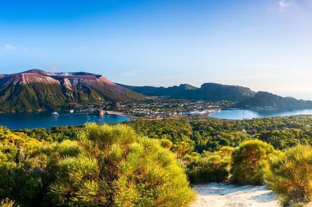 Vue aérienne d'une île volcanique montrant une ville côtière, une baie bleue avec des bateaux, et le cratère du volcan sous un ciel clair.
