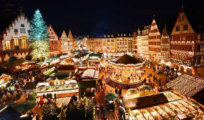 An elevated view of a bustling Christmas market at night, with a large illuminated tree, a glowing carousel, and market stalls.