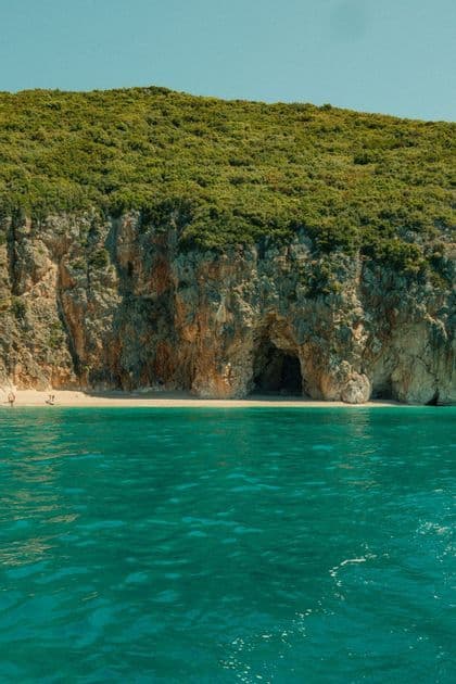 Vue depuis l'eau turquoise d'une plage de sable isolée avec une grotte marine au pied d'une falaise escarpée et arborée.
