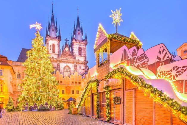 A brightly lit Christmas tree and market stalls fill a historic cobblestone square in front of a Gothic church at dusk.