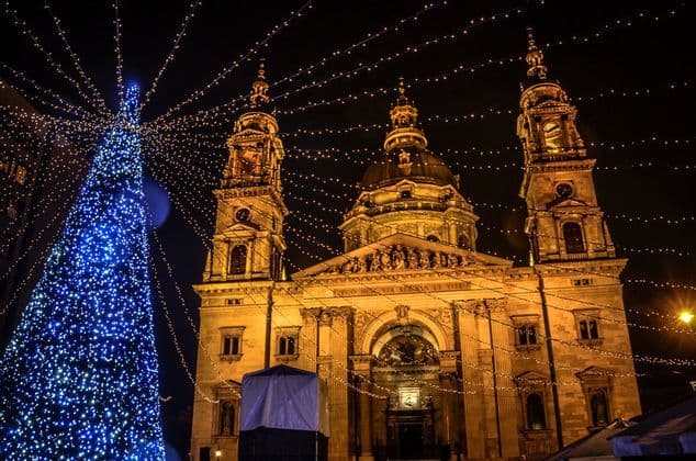 A grand cathedral at night, lit by warm lights, next to a large Christmas tree covered in blue fairy lights.
