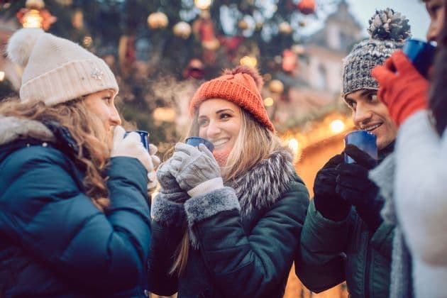 A WeRoad group trip of friends in winter hats and coats, smiling and drinking hot beverages from mugs at an outdoor market.