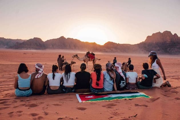 Un groupe WeRoad assis sur une dune de sable rouge, avec un drapeau jordanien, admirant le coucher de soleil sur le désert, avec des montagnes et des chameaux en arrière-plan.