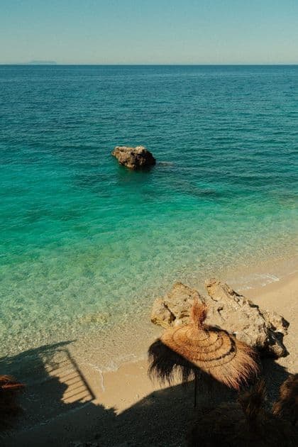 Vista aérea de una sombrilla de playa de paja en una orilla arenosa junto a aguas cristalinas de color turquesa bajo un cielo soleado.