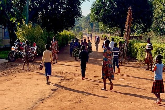 Un gruppo di adulti e bambini si allontana dalla fotocamera su un ampio sentiero sterrato, fiancheggiato da una vegetazione lussureggiante e alberi.