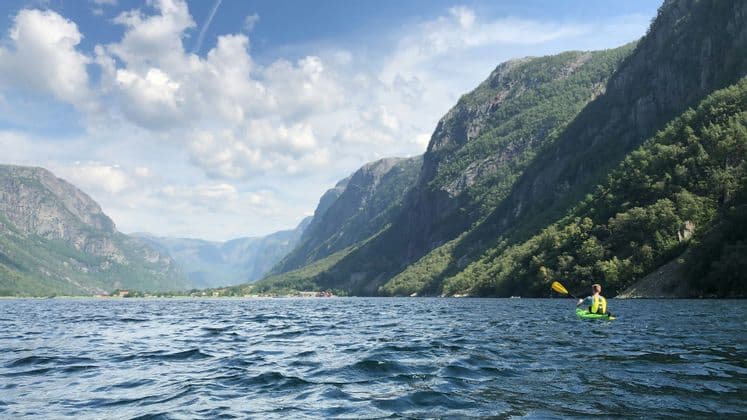 A person in a green kayak paddles on a fjord surrounded by steep, green mountains under a partly cloudy sky.