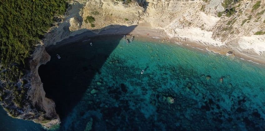 Une vue aérienne d'une crique isolée où de petits bateaux sont amarrés sur la plage et flottent dans la mer turquoise cristalline à côté de falaises abruptes.