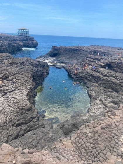 Un grupo de WeRoad nada y se relaja en una piscina natural rodeada por una costa rocosa y volcánica bajo un cielo azul claro.