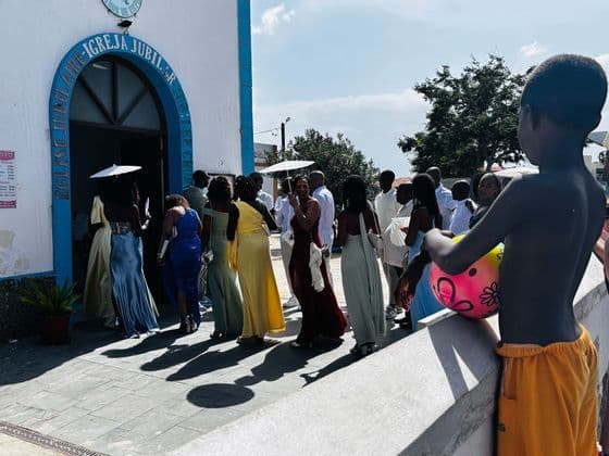 Un grupo de personas con coloridos vestidos formales sale de una iglesia, observadas por un niño que sostiene una pelota en primer plano.
